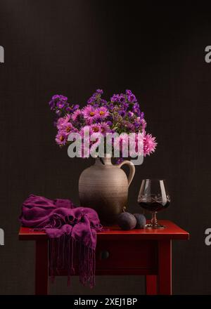 Blumenstrauß im Herbst. Viele lilafarbene Astern in einer braunen Vase. Neben einem Glas Rotwein auf einem roten Holztisch. Zwei frische Feigen. Dunkelrosa Schal. Stockfoto