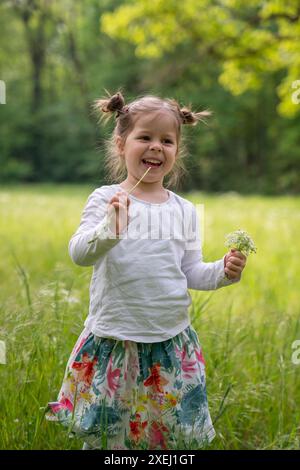 Fröhliches, kleines Mädchen spaziert durch den Park zwischen dickem, grünem Gras. Er lacht glücklich und hält Blumen an der Hand. Kindheit, Öko-Urlaub, Lachen Stockfoto