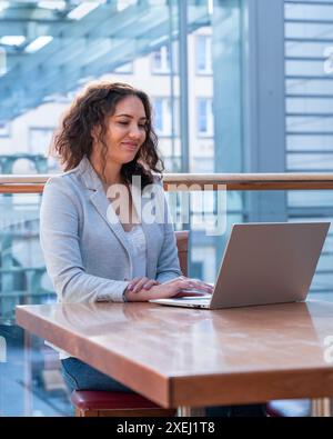 Ein Mädchen mit dunklen Haaren arbeitet an einem Tisch mit einem Laptop. Hinter einem großen Fenster Stockfoto