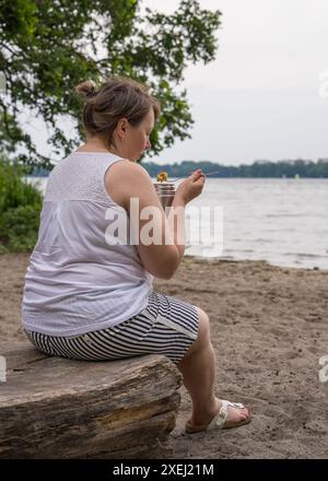 Eine übergewichtige Frau mittleren Alters ist draußen in der Nähe eines Sees und isst Nahrung aus einer Thermoskanne. Stockfoto