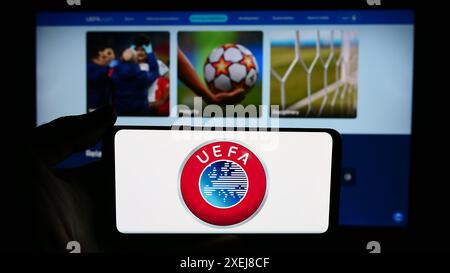 Person, die ein Mobiltelefon mit dem Logo der Union Europäischer Fußballverbände (UEFA) vor der Webseite hält. Schwerpunkt auf dem Display des Telefons. Stockfoto