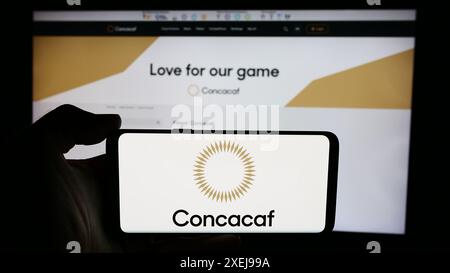 Person mit Handy mit Logo des Fußballverbands CONCACACAF vor der Webseite. Schwerpunkt auf dem Display des Telefons. Stockfoto