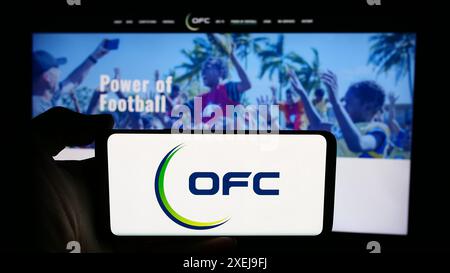 Person mit Handy mit Logo des Verbands Oceania Football Confederation (OFC) vor der Webseite. Schwerpunkt auf dem Display des Telefons. Stockfoto