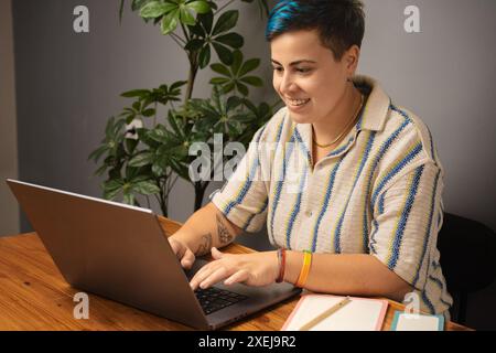 Androgyne Person mit blauem Haar, die von zu Hause aus mit einem Laptop arbeitet – fokussiert und produktiv, LGBT-Stolz-Armband, modernes Home Office-Setup, inklusive Stockfoto