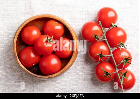 Rote Kirschtomaten in einer Holzschale auf Leinengewebe. Frische, reife Art kleiner und runder Cocktailtomaten, links noch an der Rebe. Stockfoto