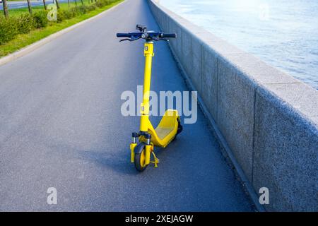 Elektrischer gelber Roller parkt auf dem Gehweg eines Asphaltweges am Ufer eines Flusses in der Stadt. Stockfoto