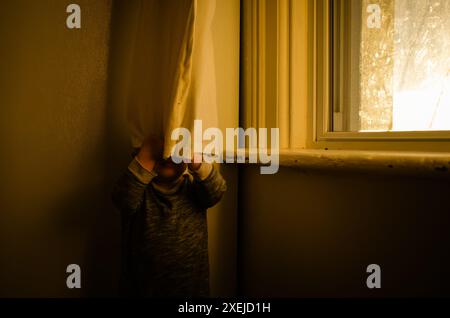 Kind versteckt sich hinter dem Vorhang in schwach beleuchtetem Raum in der Nähe des Fensters Stockfoto