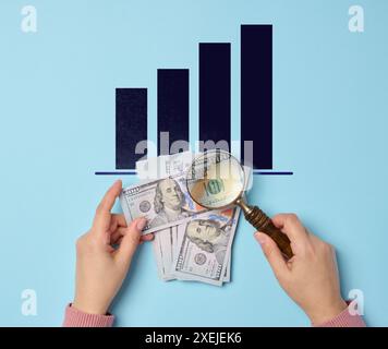100 Dollar-Scheine und eine Frauenhand, die eine Lupe auf blauem Hintergrund hält, Draufsicht Stockfoto