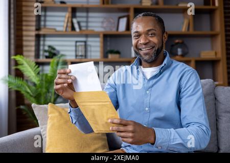 Mann öffnet glücklich den Umschlag, während er auf der Couch im gemütlichen Wohnzimmer sitzt. Happy Moment, der drinnen festgehalten wird und Freude, Begeisterung und Vorfreude ausdrückt. Stockfoto