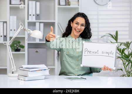 Ein begeisterter Lehrer, der während des Online-Englischunterrichts die Daumen hochlegt, während er ein Whiteboard hält, auf dem das Präsent einfach geschrieben ist. Bildungskonzept in hellem, organisiertem Büroumfeld. Stockfoto