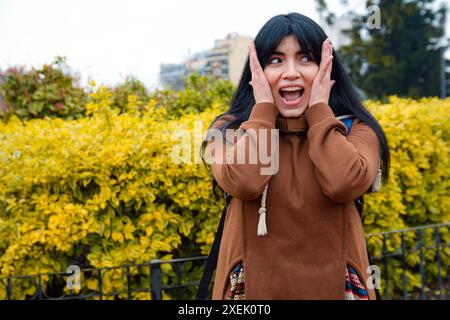Junge Lateinfrau, die im Park steht, mit den Händen auf dem Gesicht und dem offenen Mund, als Ausdruck, dass sie nicht glauben kann, was sie um sie herum sieht, Emot Stockfoto