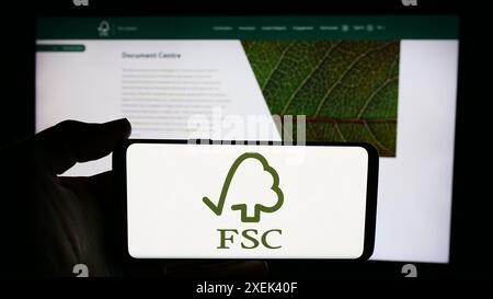 Person mit Mobiltelefon mit Logo der Organisation Forest Stewardship Council GmbH (FSC) vor der Webseite. Schwerpunkt auf dem Display des Telefons. Stockfoto