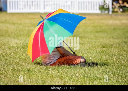 Ein Mann saß unter einem mehrfarbigen Regenschirm, auf grünem Gras, mit einem weißen Pflückzaun im Hintergrund Stockfoto