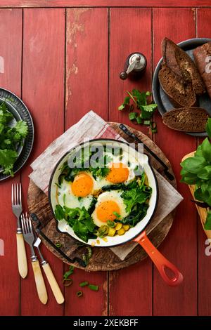 Grüne Shakshuka. Spiegeleier, Kartoffeln, Spinat und Zwiebeln in roter gusseiserner Pfanne mit Roggenbrot auf einem alten roten Vintage-Tisch Hintergrund. Traditionelle Juden Stockfoto