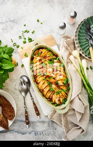 Hasselback-Kartoffel gefüllt mit Schinken, Zwiebeln, Knoblauch und Kräutern, serviert in ovaler Keramikform mit Gewürzen, Basilikum, Koriander, Roggenbrotstücken auf Grau Stockfoto