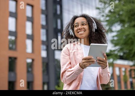 Glückliche junge Studentin mit Kopfhörern, die außerhalb des Universitätsgeländes ein Tablet verwenden. Schüler genießen Lernen und Musik in einer modernen Bildungsumgebung. Stockfoto