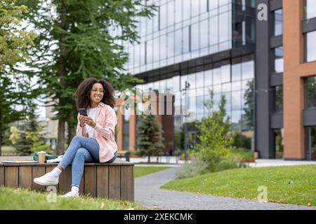 Junge Studentin, die in der Nähe des Universitätsgeländes sitzt und ein Smartphone hält. Lächelnde Frau, die legere Kleidung trägt, genießt den Außenbereich auf dem Campus mit modernen Gebäuden im Hintergrund. Entspannen und draußen lernen. Stockfoto