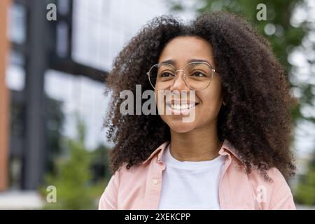 Junge Studentin mit lockigen Haaren und Brille, die in der Nähe des Universitätsgeländes selbstbewusst lächelt. Heller und positiver Ausdruck, der das Leben der Schüler und das Bildungsumfeld hervorhebt. Stockfoto