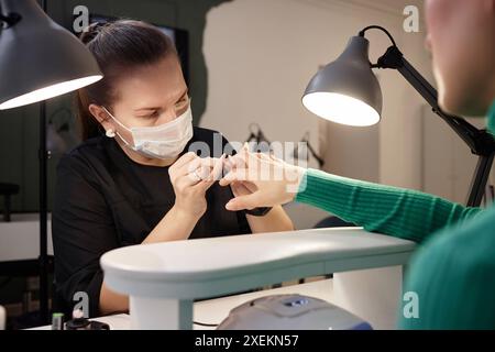 Überschulteraufnahme der professionellen weiblichen Nageltechniker, die an der Maniküre des Kunden mit Schutzmaske in einem gut ausgestatteten modernen Nagelsalon arbeitet, Kopierraum Stockfoto