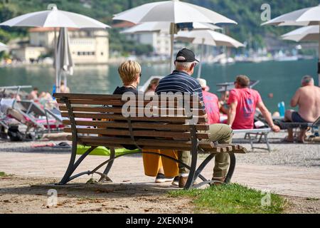 Nago Torbole, Gardasee, Italien - 27. Juni 2024: Senioren genießen eine entspannte Zeit auf einer Parkbank am Gardasee, umgeben von anderen Touristen und Strandbesuchern unter Sonnenschirmen *** Senioren genießen eine entspannte Zeit auf einer Parkbank am Seeufer am Gardasee, umgeben von anderen Touristen und Strandbesuchern unter Sonnenschirmen Stockfoto