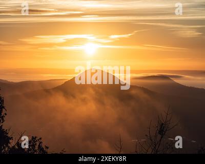 Golden Hour Sonnenaufgang über einem bewaldeten Hügel im Herbst, dicker Nebel zwischen den Bäumen, Wolkendecke und Sonnenstrahlen Stockfoto