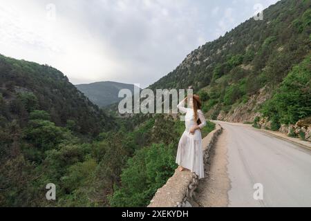 Eine Frau in einem weißen Kleid steht auf einer Straße mit Blick auf eine Bergkette. Die Szene ist ruhig und friedlich, mit dem Hut der Frau, der einen Hauch von Launen hinzufügt Stockfoto