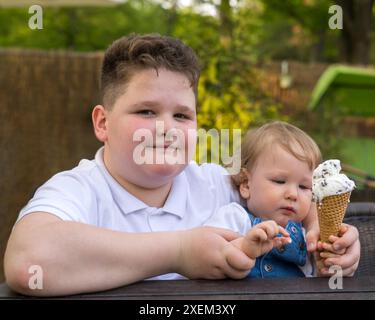 Ein glücklicher übergewichtiger Junge hält ein hellhaariges kleines Baby in den Armen, das Kind isst Eis. Brüder. Fettleibigkeit bei Kindern Stockfoto