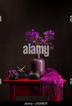 Strauß von lilafarbenen Chrysanthemen. Mehrere Zweige kleiner Herbstblumen in einer lila Vase auf einem roten Holztisch. In der Nähe ist ein großer Haufen blauer Grap Stockfoto