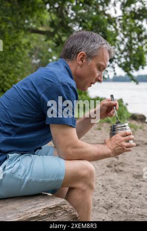 Ein Mann mittleren Alters reist, sitzt am Ufer des Sees, isst Nahrung aus einer Thermoskanne. Meditation, Genuss, Entspannung Stockfoto