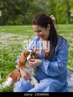 Eine schwangere Frau in einem Stadtpark sitzt auf dem Gras und hält einen süßen Cocker Spaniel Hund in ihren Armen. Liebe zu den Tieren, Stockfoto