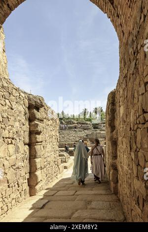 Zwei Frauen in römischer Kleidung gehen durch einen historischen Steinbogen in Merida, Spanien, mit dem antiken Amphitheater im Hintergrund. Stockfoto