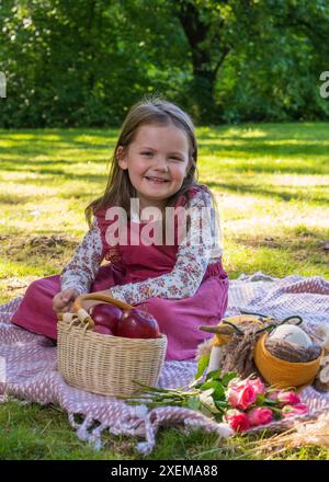 Porträt eines süßen kleinen Mädchens auf einem Picknick. Das Kind hält einen roten Apfel in der Hand und lächelt; in der Nähe befindet sich ein Obstkorb und ein Teddybär Stockfoto