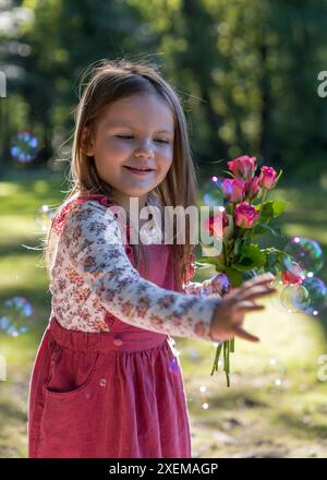 Ein kleines, süßes Mädchen in einem rosafarbenen Kleid hält Rosen in den Händen und schaut sie fröhlich an. Stockfoto