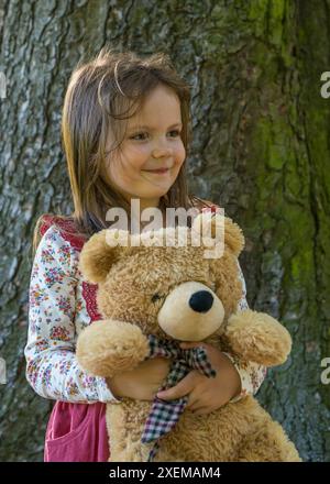 Das süße, 5-jährige Mädchen sitzt neben einem großen Baum im Park. Einen großen Teddybären halten, lächeln und in die Kamera schauen. Freundschaft, glückliches Kind Stockfoto