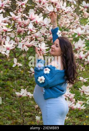 Porträt einer niedlichen schwangeren Frau im Park, gekleidet in einem blauen Kleid zwischen Magnoli-Blumen. Rückansicht. Schwangerschaft. Blühende Magnolie Stockfoto