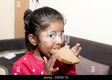 Ein junges Mädchen mit einem hellen Lächeln nimmt einen Bissen von ihrem gesunden Sandwich, während es sich in einem komfortablen Wohnzimmer entspannt Stockfoto