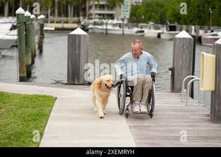 Mann im Rollstuhl mit seinem Begleithund, der auf einem Pfad am Wasser läuft; Boynton Beach, Florida, Vereinigte Staaten von Amerika Stockfoto