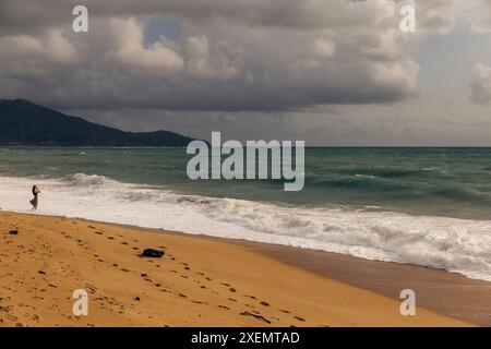 Frau stehend im Meer surfen entlang der Küste am Mai Khao Beach in North Phuket mit Blick auf den dramatischen Blick auf einen stürmischen Himmel über dem Meer bei s... Stockfoto