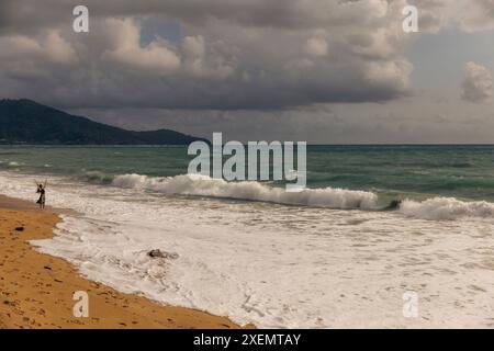 Frau, die am Ufer steht, mit ihren Armen in der Luft, und beim Sonnenuntergang auf das Meer unter einem stürmischen Himmel am Mai Khao Beach in North Phuket blickt Stockfoto