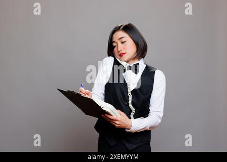 Junge asiatische Kellnerin in Uniform, die auf einer Zwischenablage steht, während sie im Festnetz telefoniert. Junger Rezeptionist im Restaurant plauderte mit dem Manager am Telefon und machte sich Notizen Stockfoto