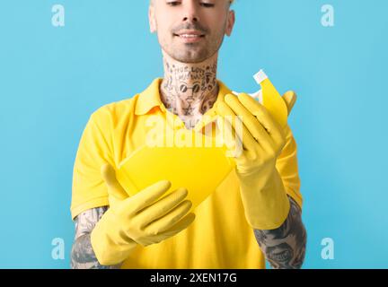 Junger tätowierter Mann mit Sprühflasche Waschmittel auf blauem Hintergrund, Nahaufnahme Stockfoto