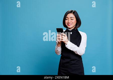 Hotelrezeptionist hält ein Smartphone und überprüft die Nachricht zufriedener Gäste. Asiatische Kellnerin mit aufgeregtem Gesichtsausdruck, die den Service mit positiven Bewertungen auf dem Handy liest Stockfoto