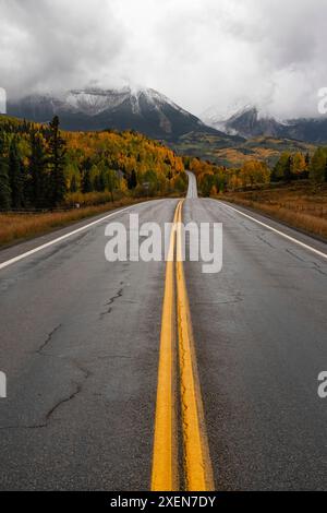 Eine asphaltierte Straße führt in die Ferne, in Richtung der malerischen Aussicht auf herbstfarbene Bäume mit grauen Wolken über den schneebedeckten Berggipfeln Stockfoto