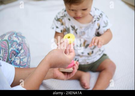 Netter Junge, der mit der Pyramide spielt, runde Kreise stapelt, mit seiner Mutter, die ihn überwacht, zusammen auf der Leinendecke sitzt. Das Konzept Stockfoto