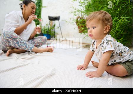 Niedlicher Junge, 10 Monate alt, in khakifarbenen Shorts und weißem Hemd mit naturgemustertem Muster, sitzend auf einer Decke auf dem Boden im Garten. Seine Mutter ist b Stockfoto