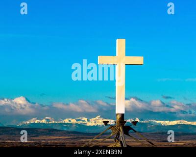 Nahaufnahme eines weißen Kreuzes eines Kirchturms mit blauem Himmel, Wolken und schneebedeckten Gebirgszügen im Hintergrund; Calgary, Alberta, Kanada Stockfoto