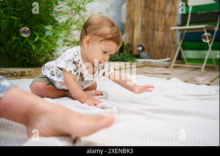 Süßer Junge, der mit Seifenblasen auf der Terrasse im Hinterhof spielt. Rothaariges weißes Kind fängt Blasen im Freien und zeigt Interesse an fliegenden Blasen. Kind Stockfoto