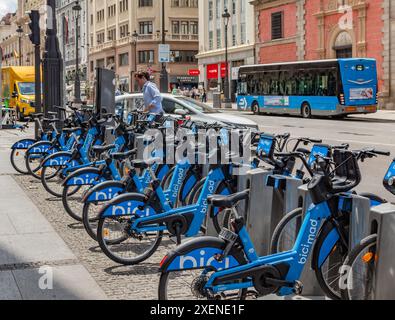 06.19.2024, Madrid, Spanien: Mann in blauem Hemd parkt ein BiciMAD-Elektrofahrrad an einer Dockingstation in einer Straße mit Bus und Verkehr Stockfoto