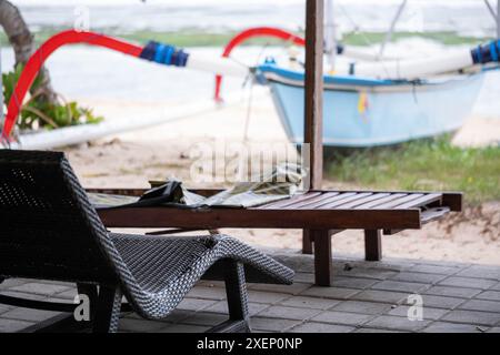 Eine entspannende Strandszene mit einem Loungesessel und einer Holzbank auf einer gefliesten Terrasse. Im Hintergrund ist ein traditionelles Auslegerkanu zu sehen Stockfoto