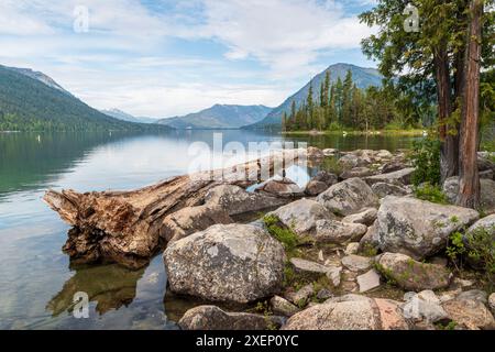 Felsen, Felsbrocken und ein umgestürzter Baum säumen die Ufer des Lake Wenatchee, mit den Kaskadengebirgen in der Ferne. Stockfoto